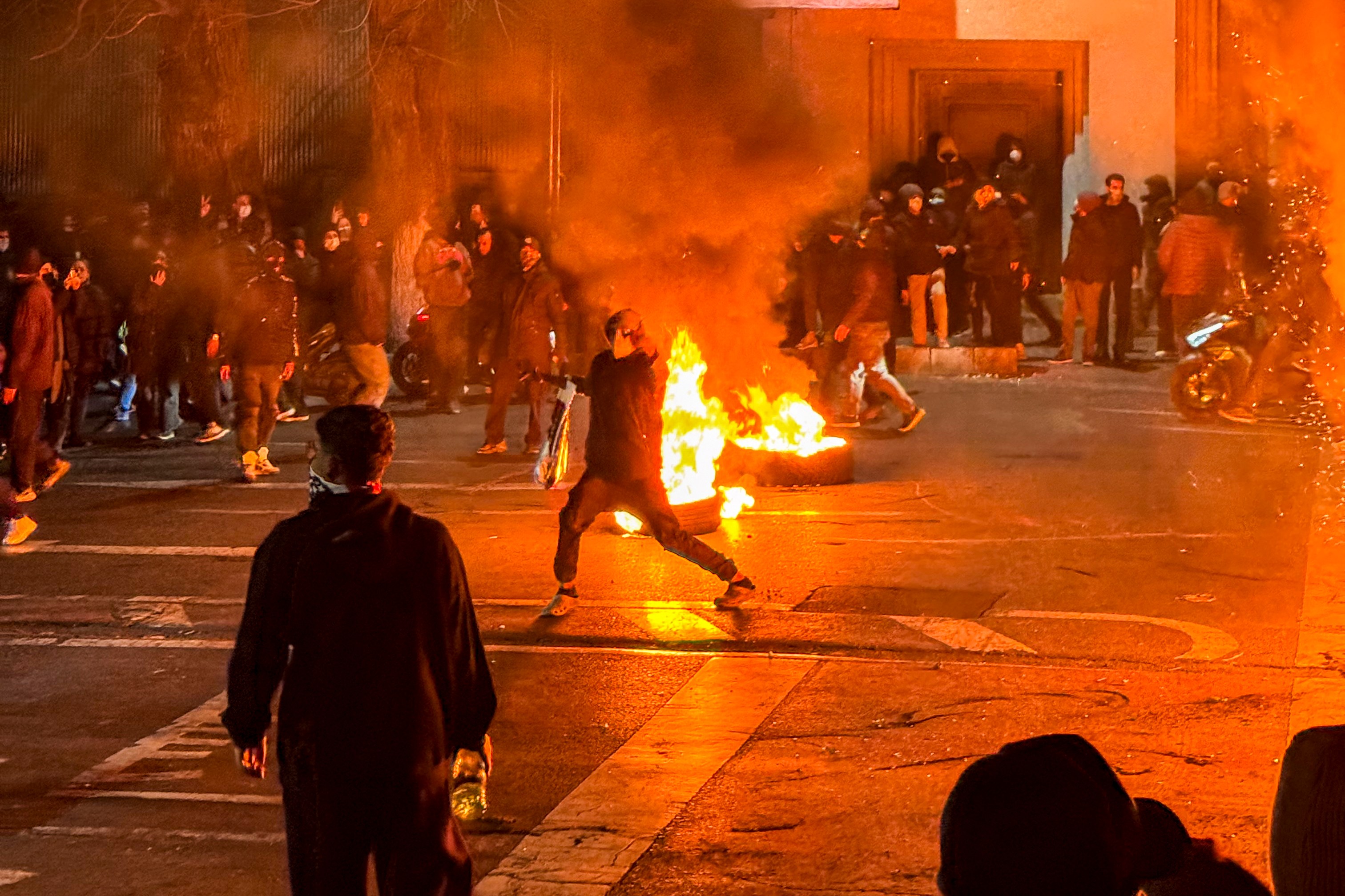 Iranians protesting in Tehran block a street with a tire fire, surrounded by smoke and other protestors.