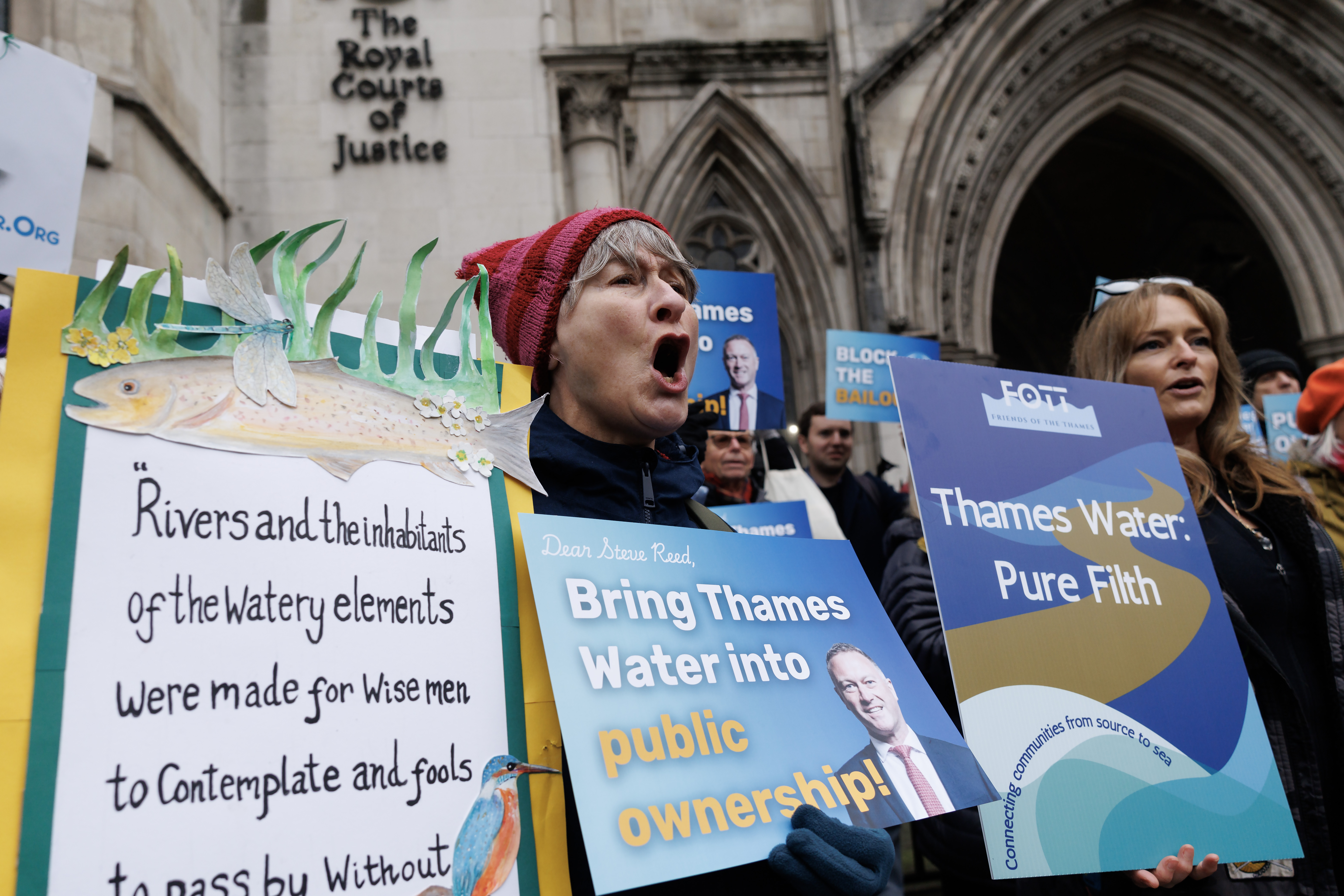 A woman in a red hat shouts while holding a sign about rivers and the watery elements, and other protestors hold signs saying "Bring Thames Water into public ownership" and "Thames Water: Pure Filth" outside the Royal Courts of Justice.