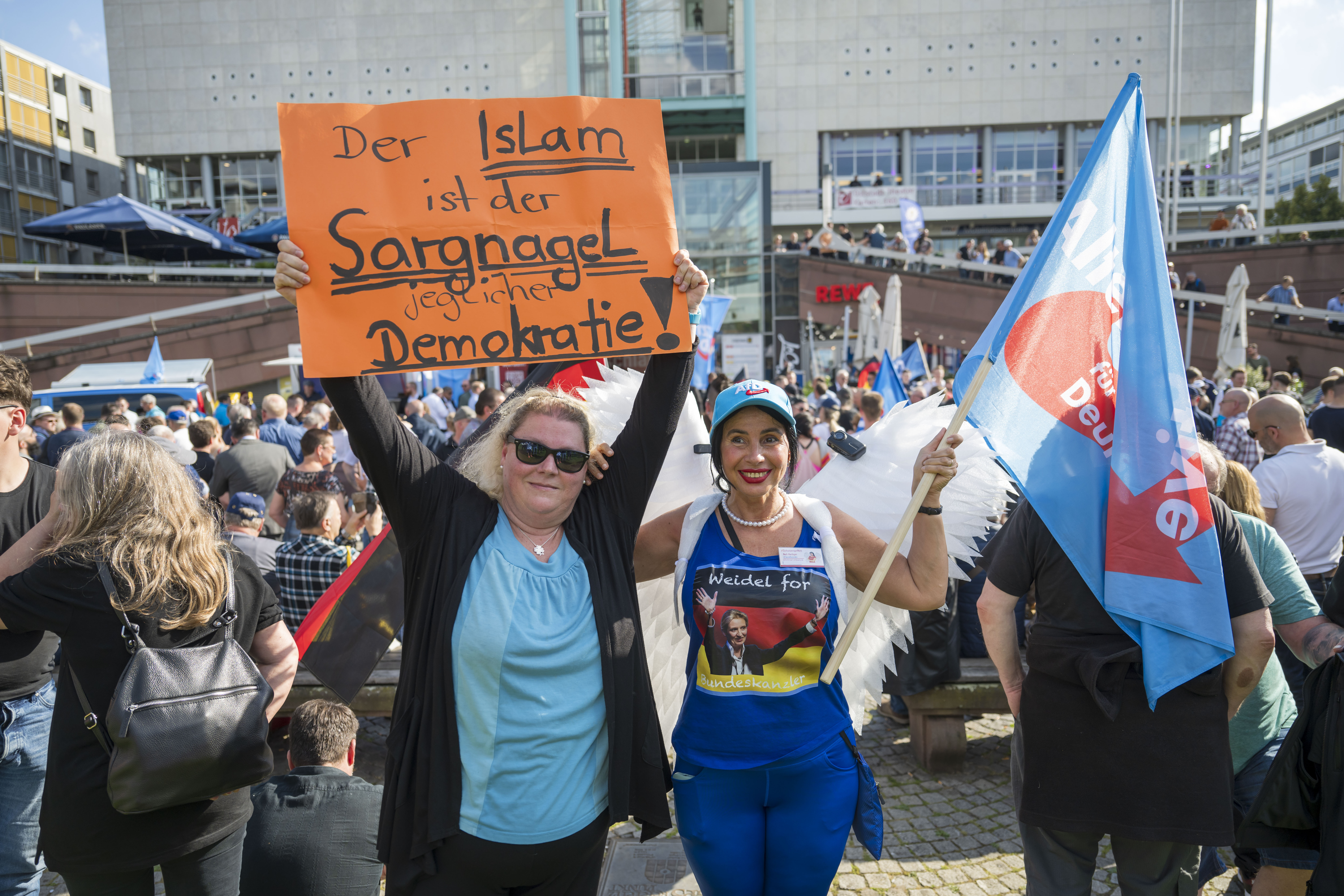 Two women at an AfD protest, one holding a sign that reads "Islam is the nail in the coffin of democracy" and the other wearing a "Weidel for Chancellor" shirt with angel wings.