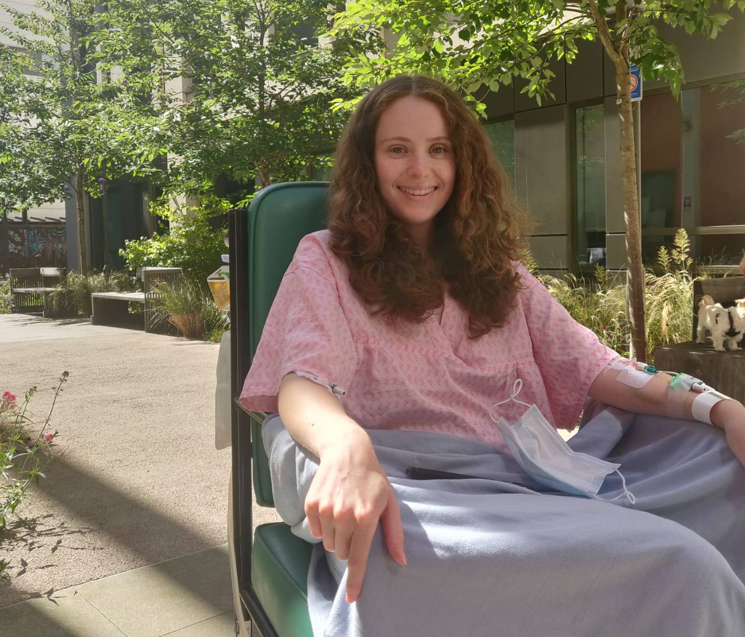 Holly Masters smiling in a wheelchair outdoors while wearing a hospital gown and having an IV in her arm.