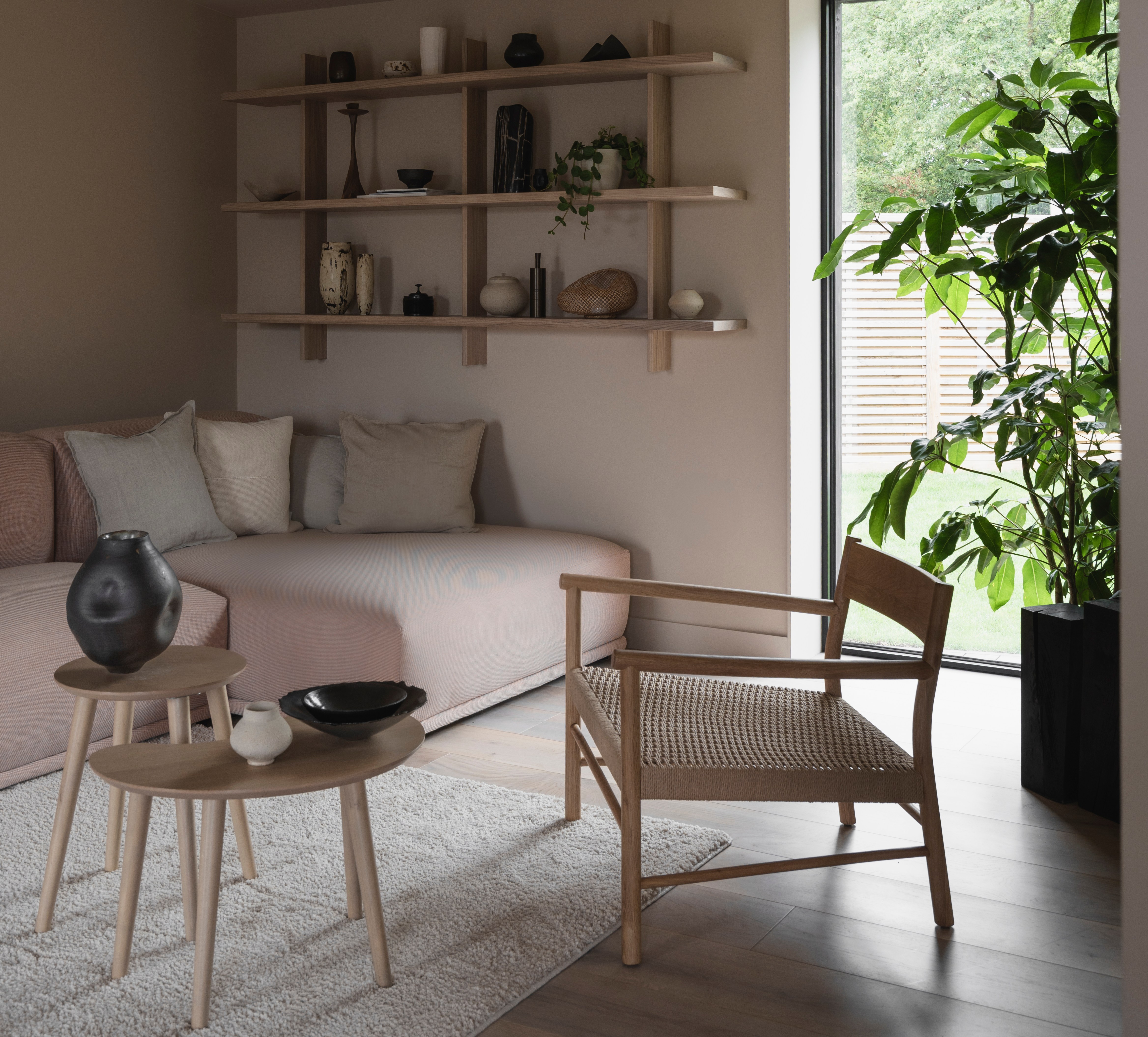Living room with light pink couch, wooden shelving unit, woven chair, and large window.