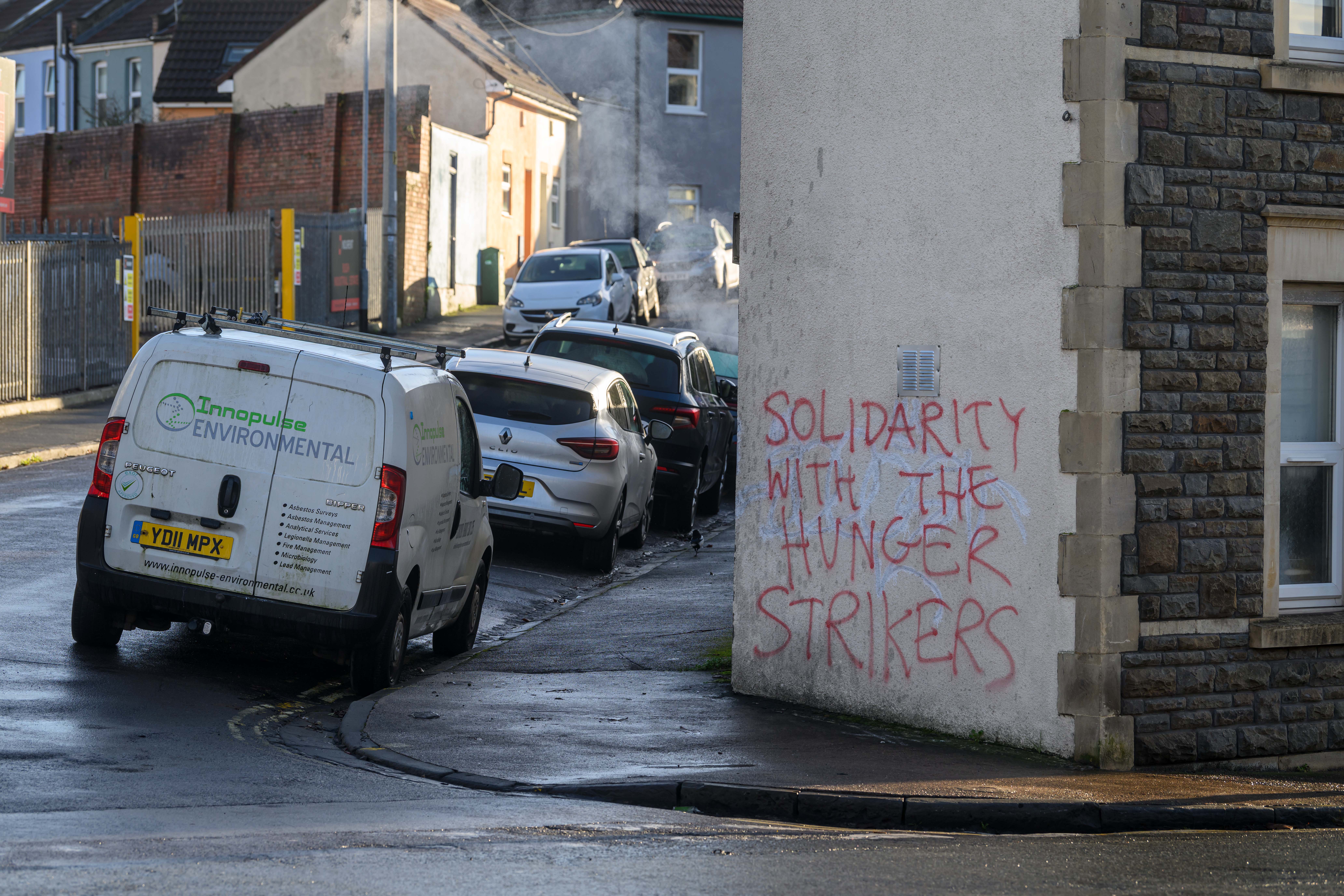 A street scene with cars parked along a curb and graffiti on a white building that reads "SOLIDARITY WITH THE HUNGER STRIKERS".