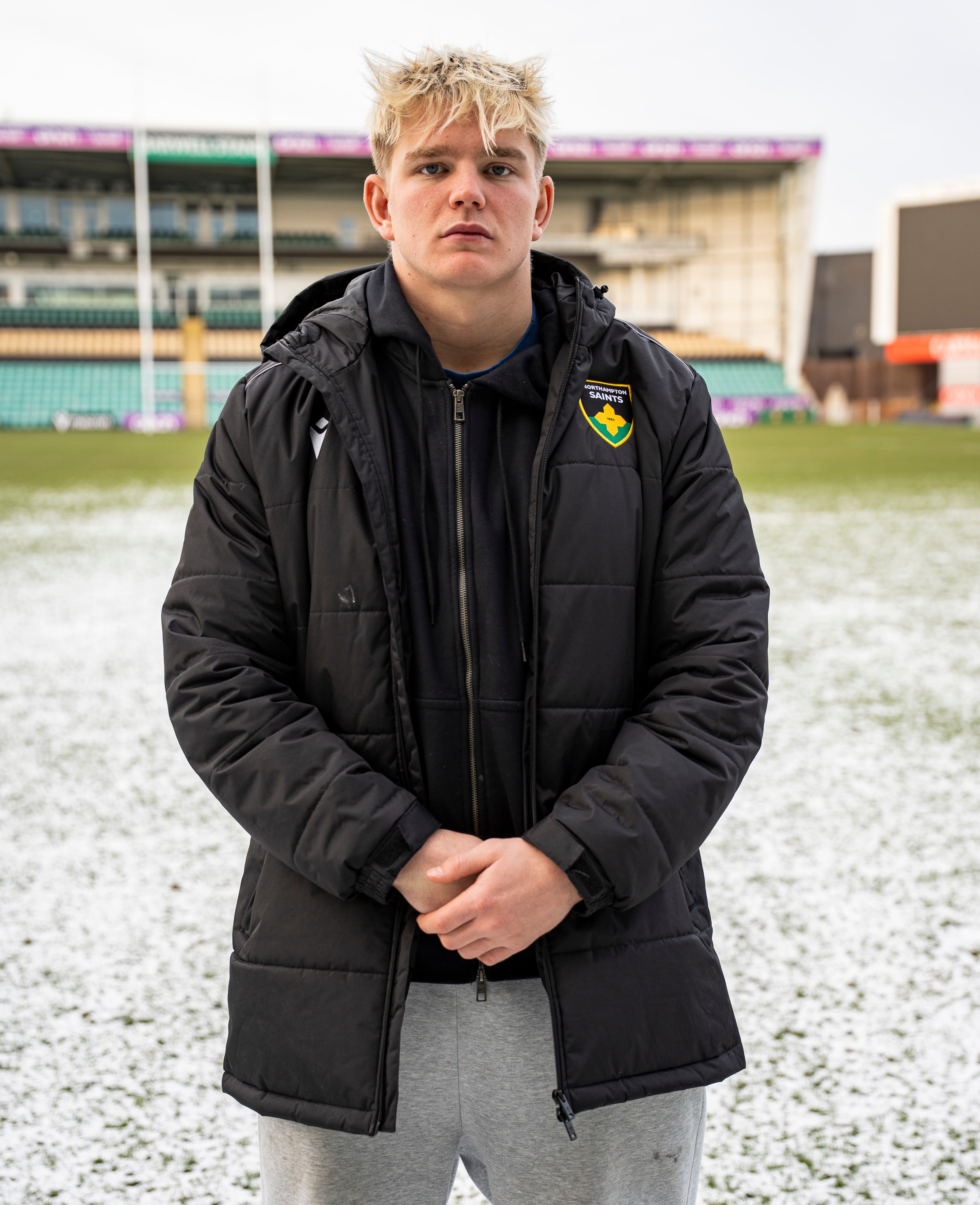 Henry Pollock (20) in a black jacket with a Northampton Saints emblem, standing on a snow-dusted field at Franklin's Gardens.