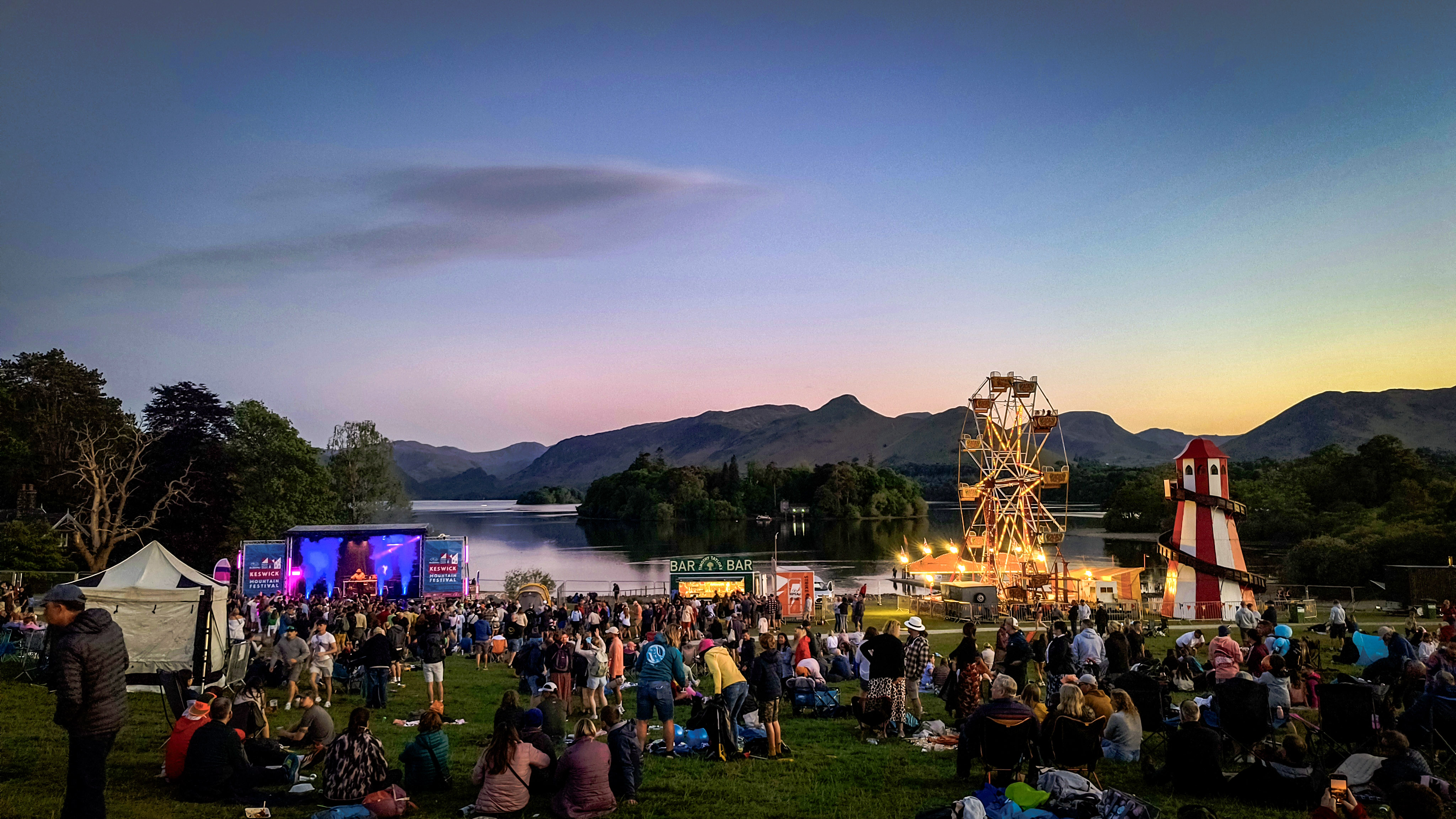 Crowds at the Keswick Mountain Festival with a Ferris wheel, lake, and mountains in the background at sunset.