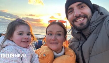 A woman and her husband pose with their young daughter, all wearing coats, at the seaside.