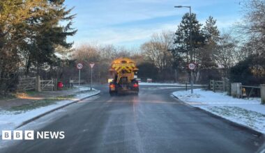 The back of a yellow gritter lorry, with a yellow and red striped sign that says "Spreading". It is at a T-junction, with the verges mostly covered in snow.