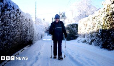A woman walks through a snowy lane wearing a wooly hat, using poles.