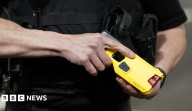 Stock image of an officer demonstrating a new Taser. It is a close up shot of a man's hands holding a yellow taser, with the name Taser 7 on it and there are two red cartridges visible.