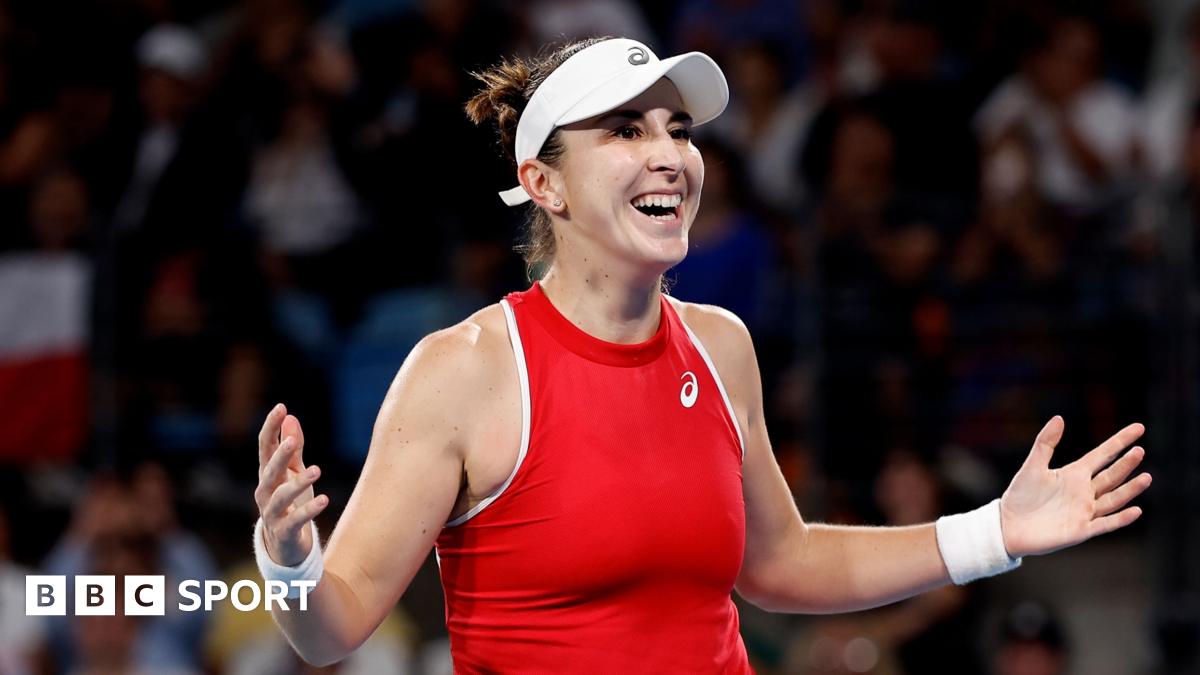 Belinda Bencic, in red tennis top and white visor, celebrates with a smile and arms spread wide, with crowd visible behind her