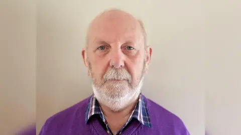 BBC Wilbert is mostly bald, with some hair on the sides of his head and a few wispy strands on the top. His grey beard is uniformly short, apart from his moustache , which is slightly longer. He wears a purple v-neck sweater over a checked shirt. 