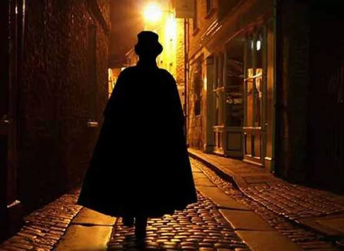 Colour photograph of the silhouette of a man walking away from the camera down a cobbled street. He wears a long cape and top hat. A light shines above a shop front ahead of him.