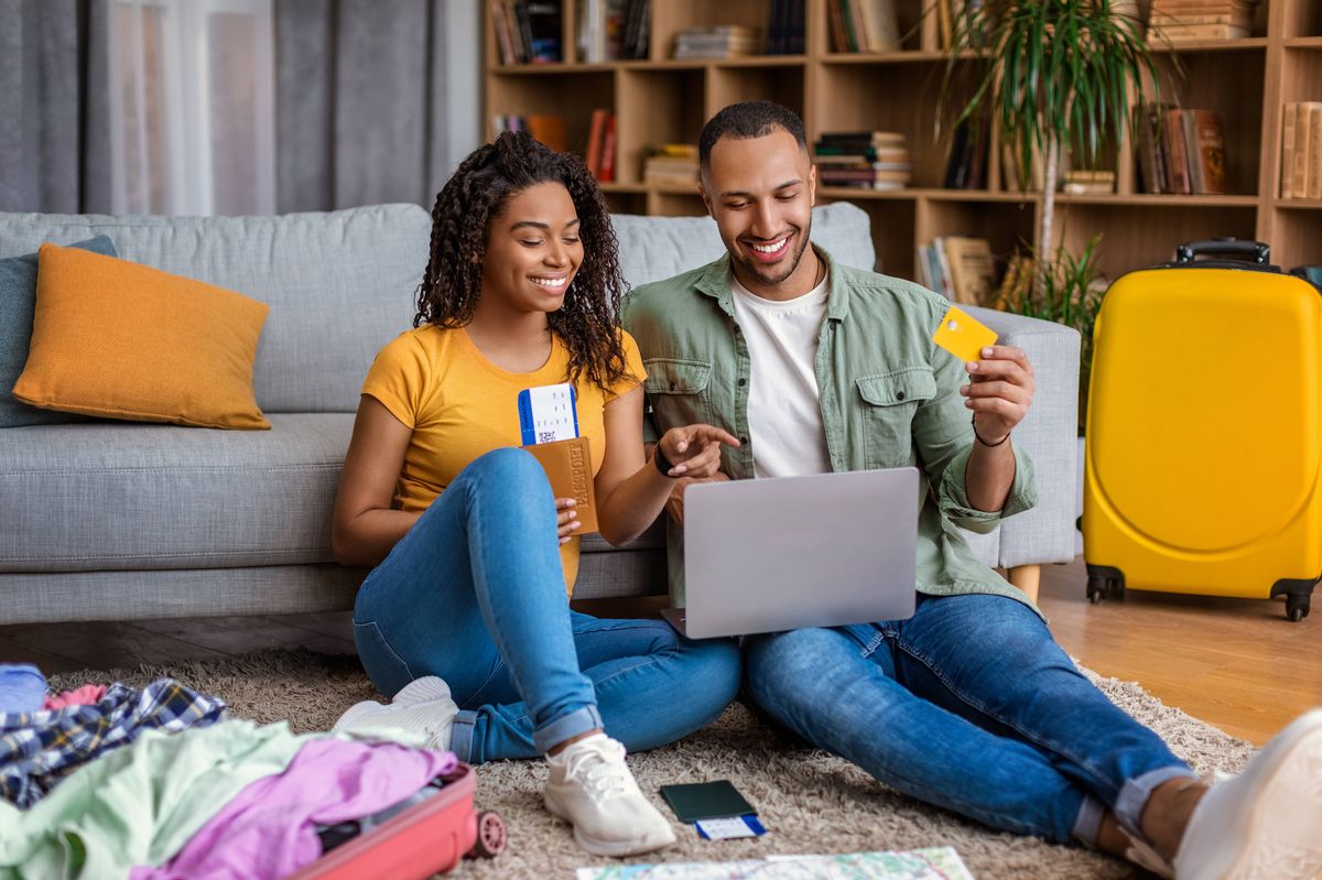 Excited black spouses using laptop and booking hotel online with credit card, lady holding passports with tickets, couple sitting with suitcases on floor at home