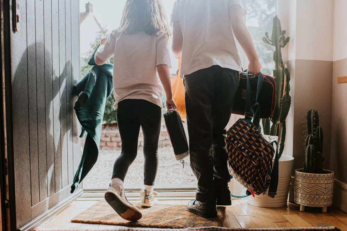 Two young children exit a doorway at the same time, holding backpacks and lunch bags and wearing a school uniform. Image depicts a morning and the school day ahead.
