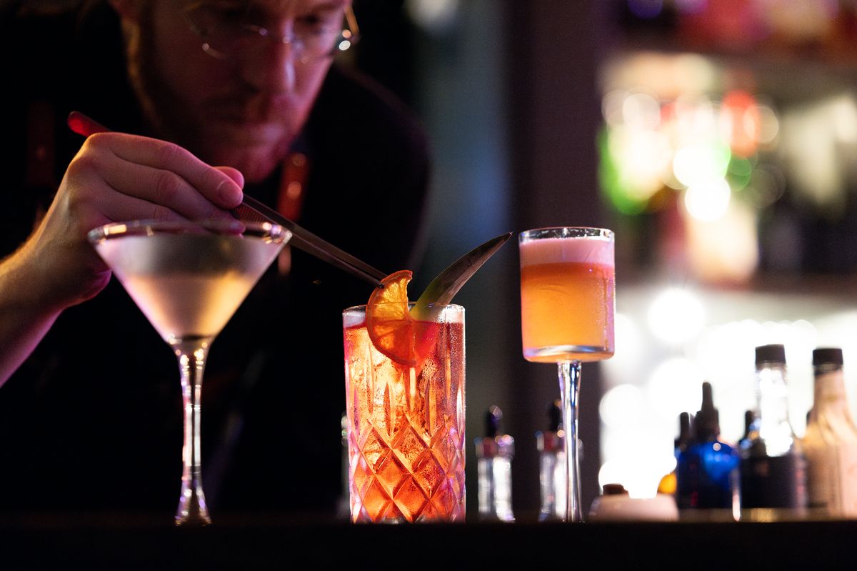 A still photograph of a bartender as he puts an orange slice into a cocktail. The drinks are vibrant and brightly lit, standing out against the low-key lighting of the background. The drinks and bartender's hand are in focus, while the rest remains blurry.