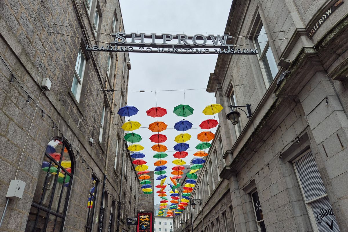 Aberdeen, Scotland, UK - July 13, 2024: A street view in the downtown of Aberdeen. Two people walking along the buildings. Buildings made of granite - grey colour. Above, the Umbrella Project, about Neurodiversity, in Shiprow, a historic street near Aberdeen Harbour, Scotland