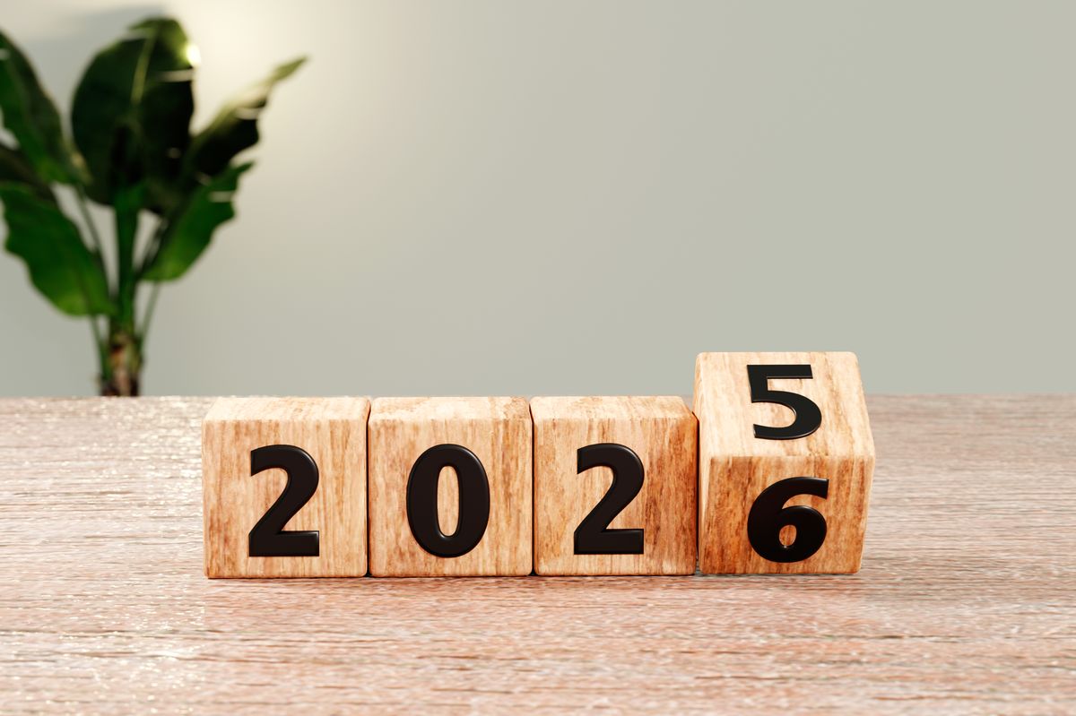 3D render of wooden cubes displaying 2026 on a tabletop, indoor scene with a blurred green plant in the background. No people. Neutral, minimal style with copy space.