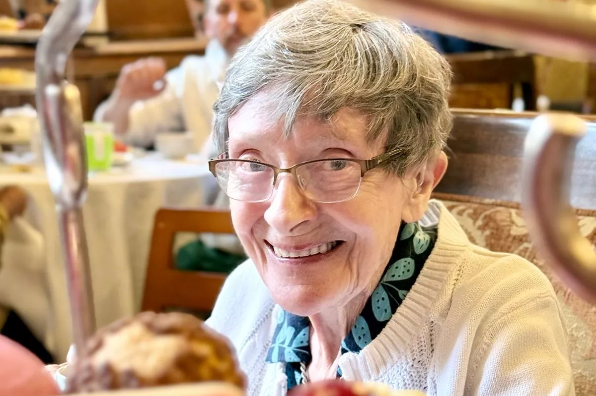 Iris Morgan, an elderly lady, looks at the camera and is smiling. She has grey hair, dark rimmed glasses, and fair skin