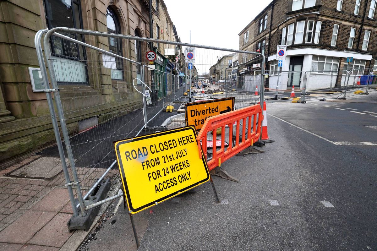 An urban street is depicted with barricades and warning signs indicating ongoing roadworks. The road is partially blocked, and caution is advised for pedestrians and vehicles. The surrounding area includes buildings and street infrastructure.