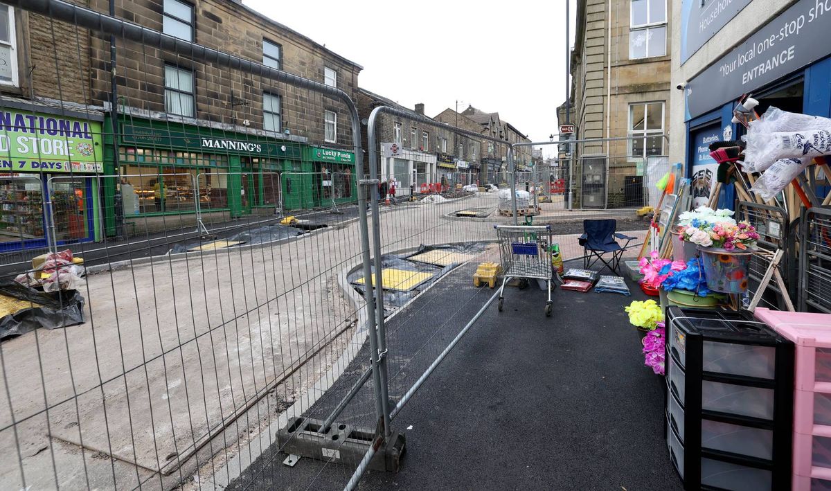 An urban street scene with a newly paved road, temporary fencing on the left side, and a shop displaying various items on the right. The background features several buildings, and the overall area appears to be undergoing some construction or renovation activities.