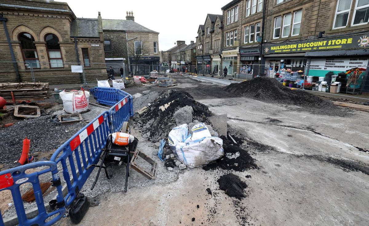 An urban construction site with visible infrastructure work, including barriers, debris, and machinery. Buildings and shops are visible in the background.