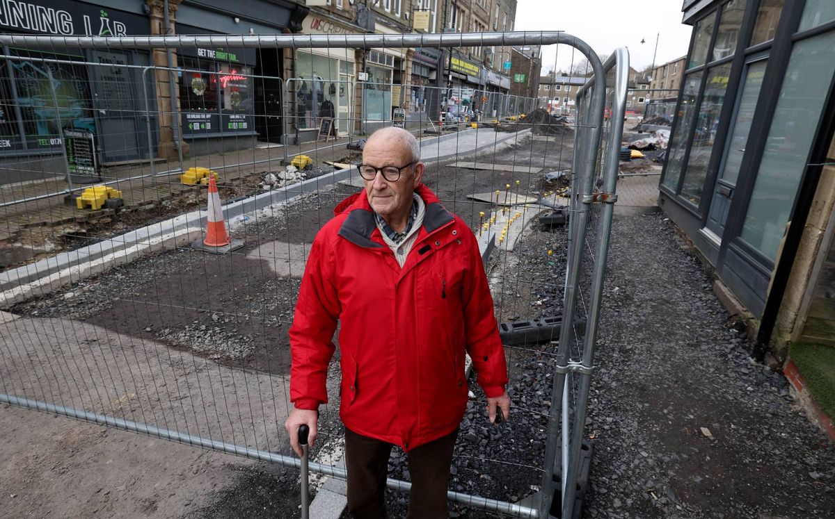 An individual dressed in a red jacket is standing near a metal barrier, presumably on a street under construction, with buildings visible in the background.