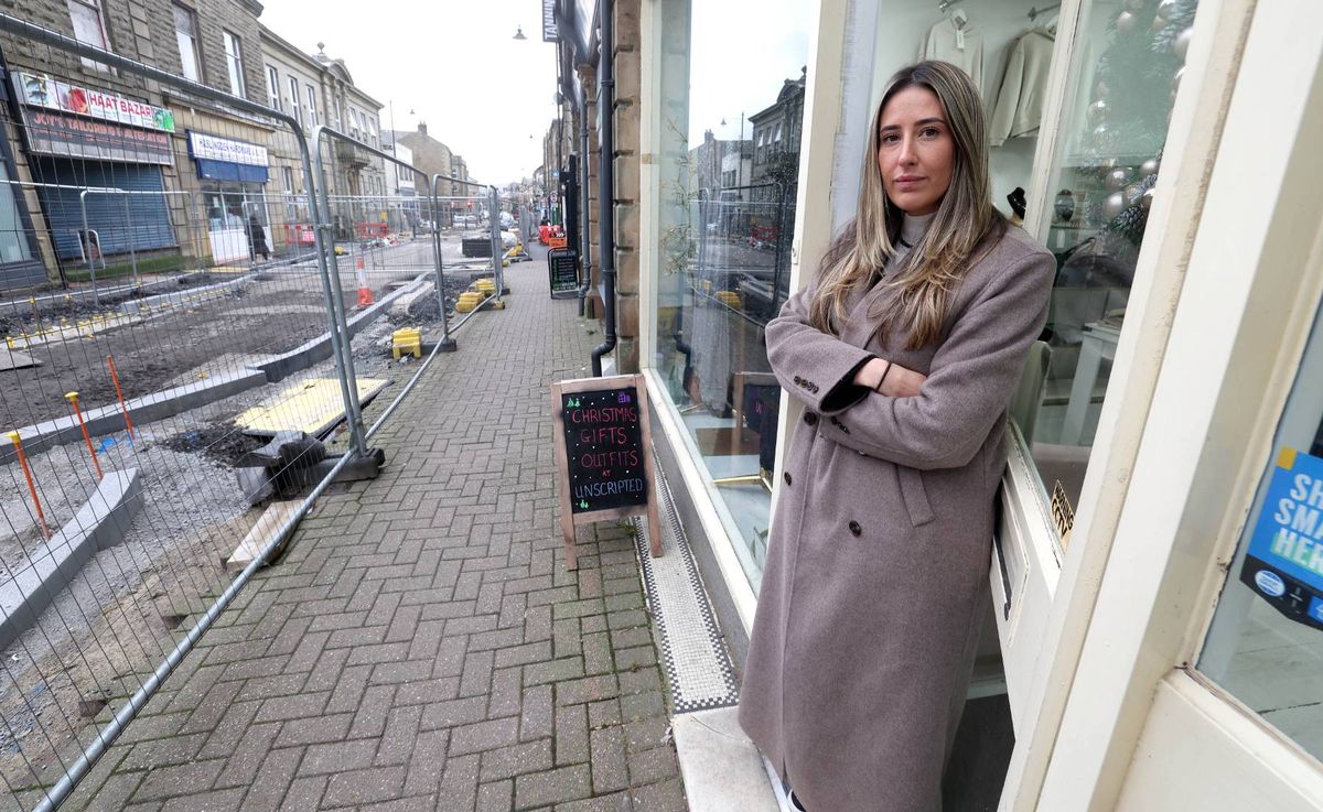 An individual dressed in a long overcoat stands on a sidewalk adjacent to a storefront, with construction barriers and an urban streetscape in the background.