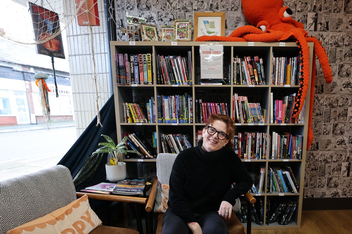 Owner Cathy Charnley inside Kraken Comics. She is dressed in black and sat on an armchair in front of a bookcase.