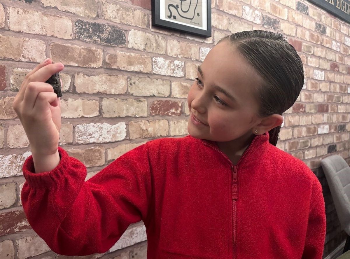 A young girl around nine years old, holding up a rock - looking at it thoughtfully. She has dark hair in a ponytail and a red zip up fleece.