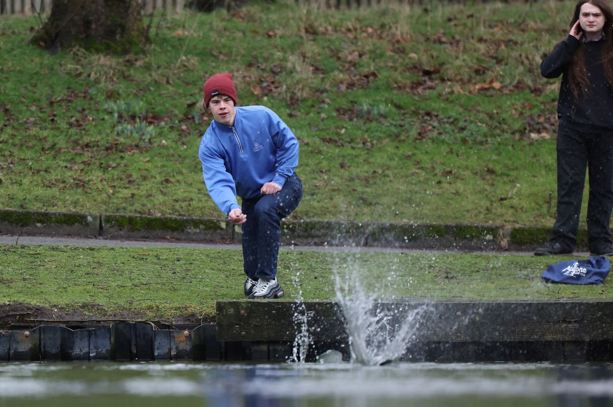 A man in a blue jacket and red hat skims a stone on a lake