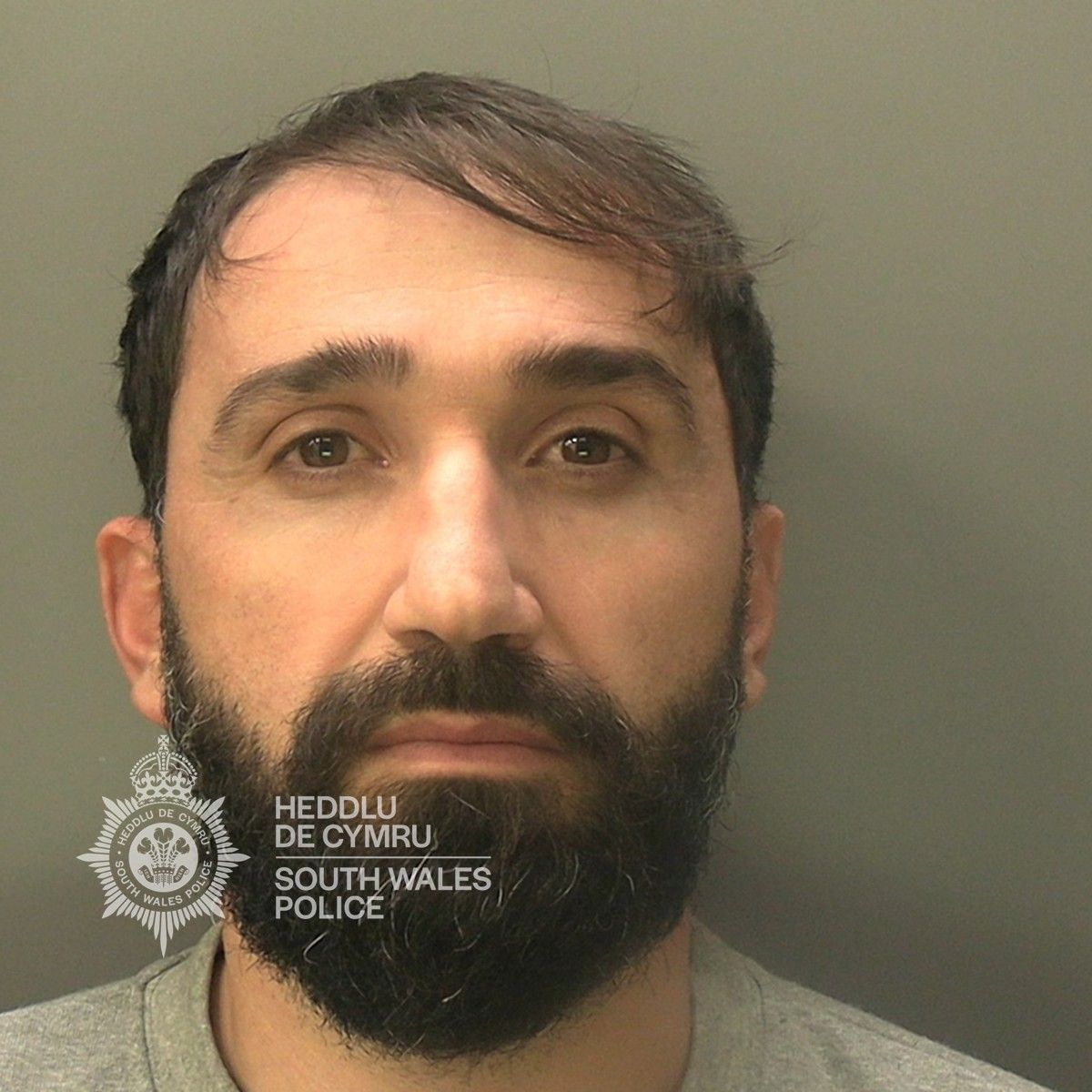 Custody photo of Kushi Alket, 35, who has black hair, a black beard and is wearing a grey tracksuit.
