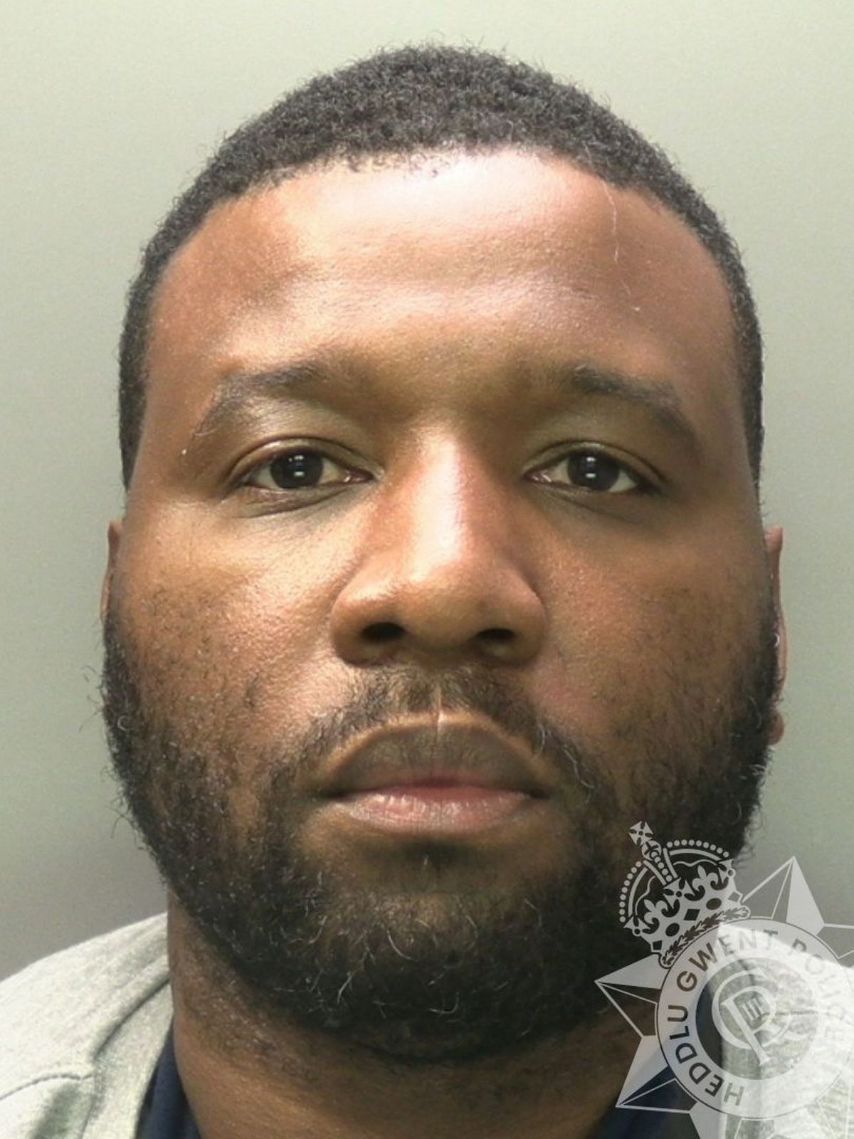 Custody photo of Michaek Dumbuya, 37, who has short black hair, a black beard, and is wearing a grey jacket and a navy t-shirt.