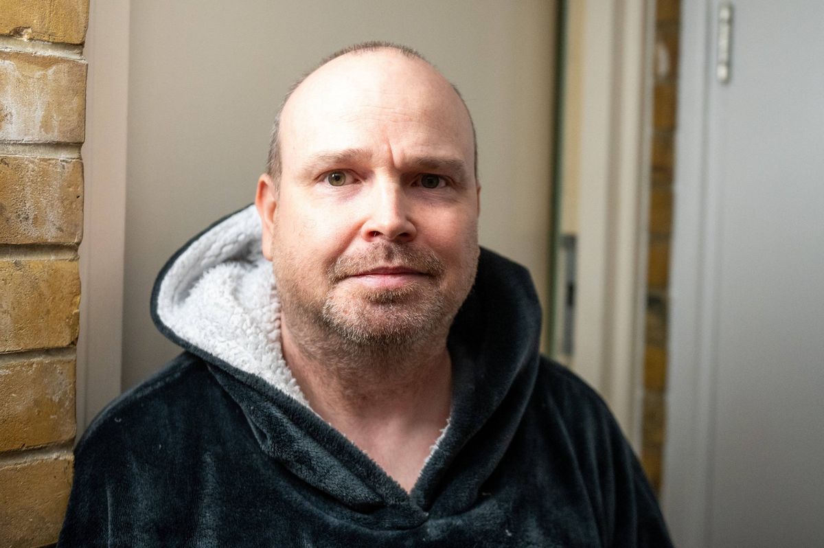 A man wearing a black hooded jacket, is standing in front of a brick wall, with a white door partially visible in the background.