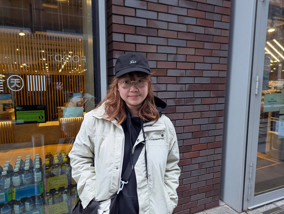 An individual is standing in front of a brick building, dressed in a white jacket and a black cap. The building features a glass storefront showcasing a variety of products.