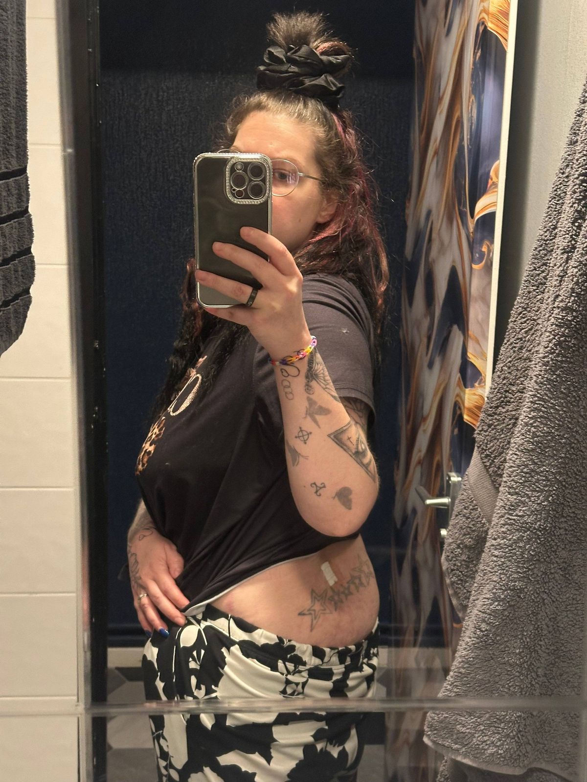 An individual with visible tattoos on their arms is seen taking a selfie in front of a mirror. The background reveals a room with a glass door, and a towel is hanging on the adjacent wall.