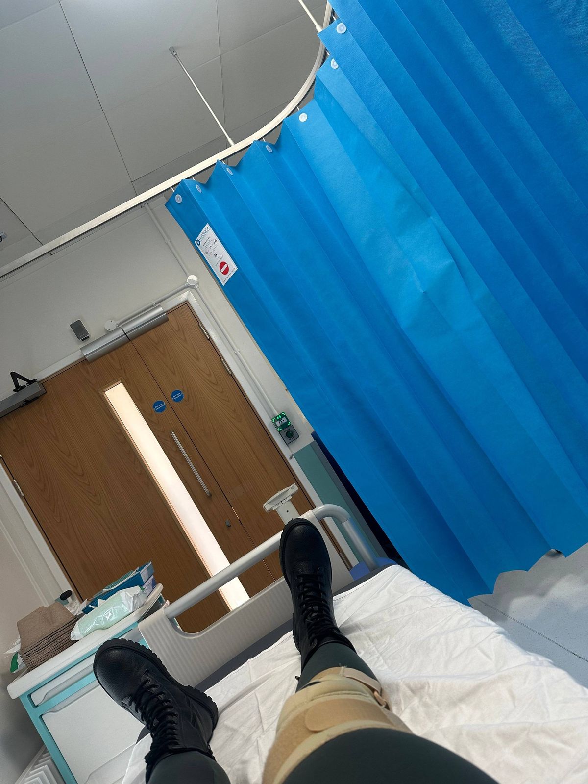 In a hospital room, an individual is resting on a hospital bed, dressed in dark trousers and black boots. The room is equipped with blue curtains, a wooden door, and various medical equipment. The setting suggests a healthcare environment.