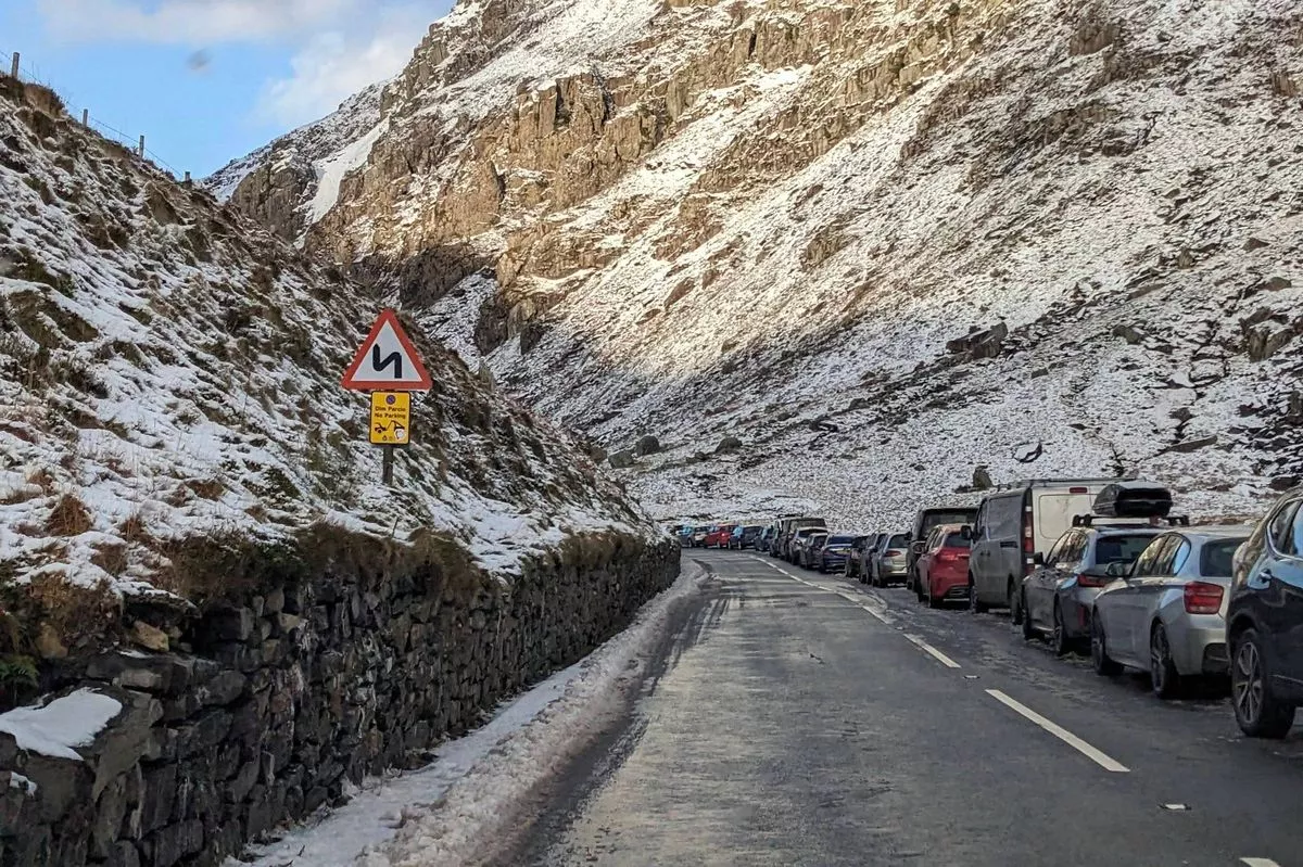 A mountainous road covered in snow, with vehicles parked along the side, indicating potential traffic congestion or a scenic winter landscape.