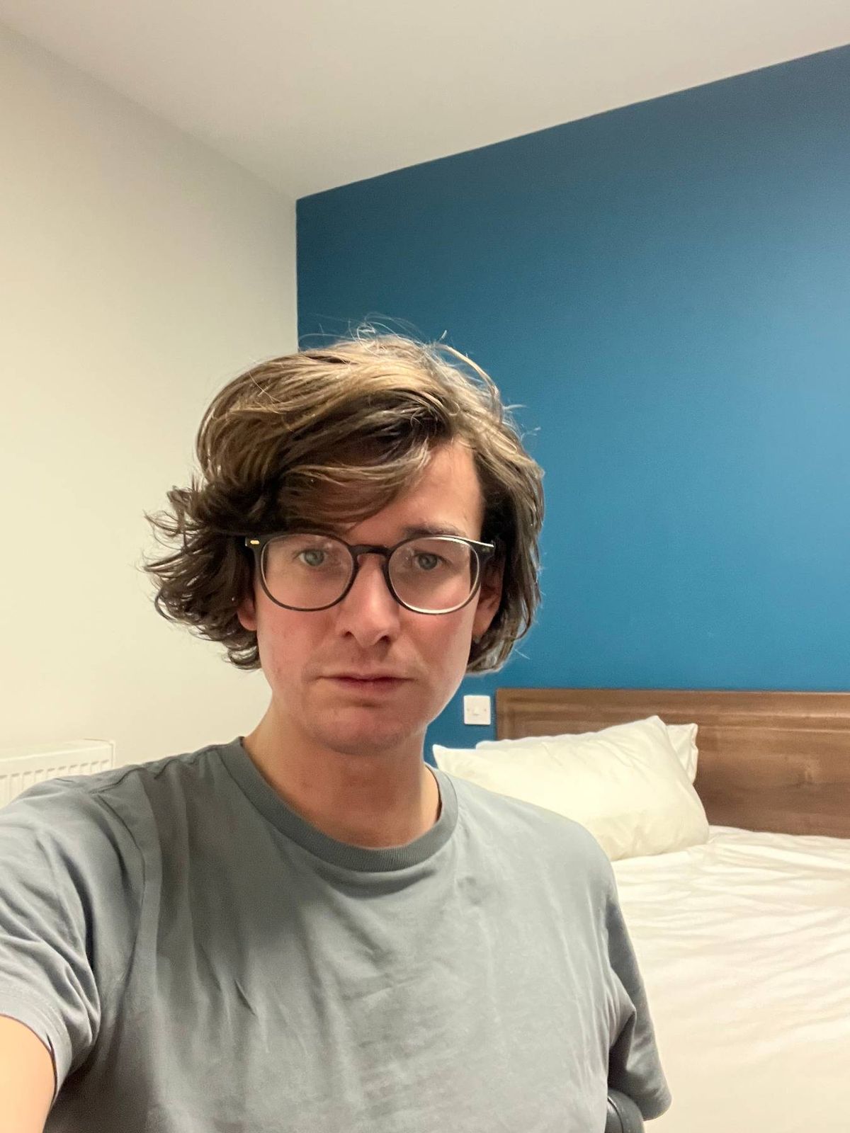 An individual with short, curly hair and wearing glasses stands in front of a blue wall. In the background, a bed with white pillows and a wooden headboard is partially visible. The person is dressed in a gray t-shirt and is looking directly at the camera.