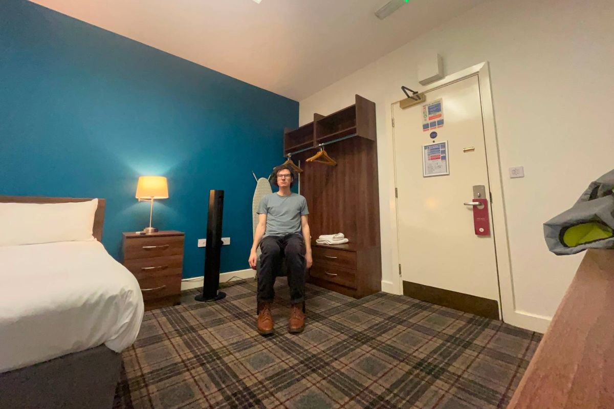 An individual is standing in a bedroom, which features blue walls, a carpeted floor, and a white bed. A lamp is placed on a nightstand, and there is a wooden cabinet with an extinguisher attached to the wall. The room has a warm and cozy atmosphere.