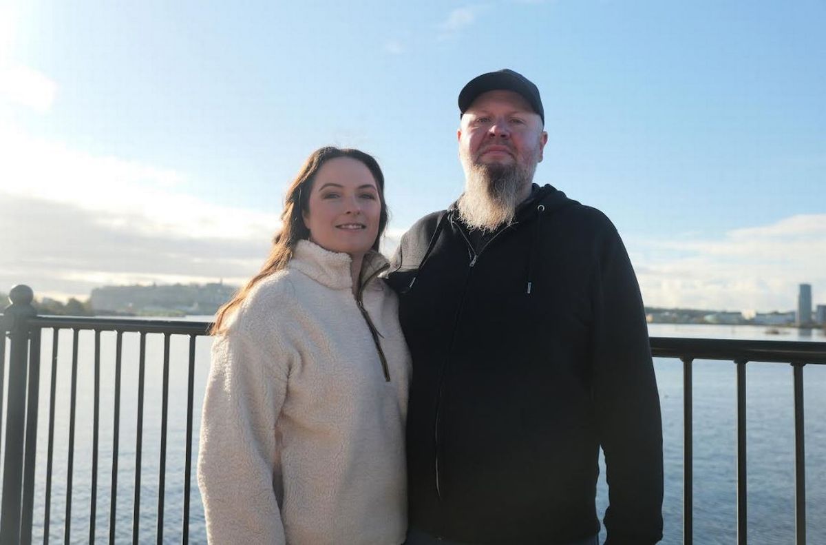 A woman in a cream fleece and a man dressed in black with a cap and a beard smiling 