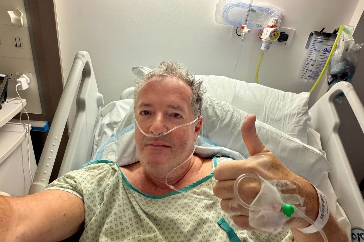 An individual, likely a patient, rests in a hospital bed, wearing a hospital gown. They have an oxygen tube attached to their nose and are making a thumbs-up gesture with one hand. The setting is a hospital room, evident from the medical equipment and the bed's design.