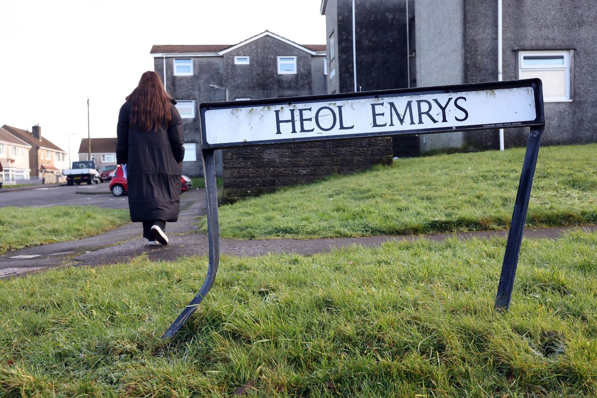 A young woman with long dark hair and an ankle length black coat walking, a sign that reads Heol Emrys in the forefront