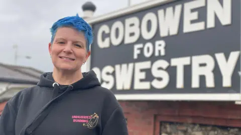 BBC A blue-haired woman, wearing a black hoodie, standing in front of a black and white railway sign, which reads, "Gobowen for Oswestry".