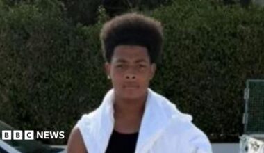 Maliki stands in front of the camera with a serious expression. He is wearing black shorts, a black vest and a white top draped across his shoulders. He appears to be on a street as cars can be seen in the background. It appears to be warm and sunny weather.