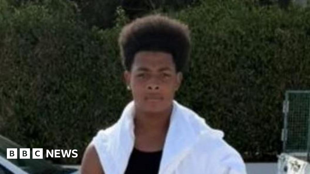 Maliki stands in front of the camera with a serious expression. He is wearing black shorts, a black vest and a white top draped across his shoulders. He appears to be on a street as cars can be seen in the background. It appears to be warm and sunny weather.