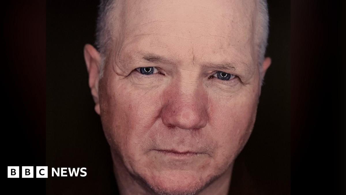 Finbar Polin is staring intently at the camera. It is a close up of his face against a black background. He has short grey hair and blue eyes.