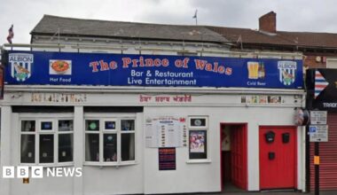 A white building with bay windows and a red door with The Prince of Wales in red writing on a blue sign. There are two bay windows, to the left of the door, with the exterior of the building painted white.