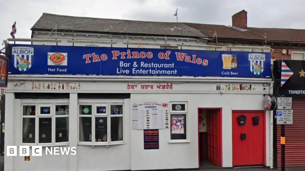 A white building with bay windows and a red door with The Prince of Wales in red writing on a blue sign. There are two bay windows, to the left of the door, with the exterior of the building painted white.