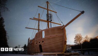 Millie Bobby Brown and David Harbour in character as Eleven and Jim Hopper. Eleven wears red shorts over grey leggings and a grey jumper. Jim Hopper wears a black leather jacket, a black beanie hat and dark trousers.They are speaking to director Frank Darabont on the set of Stranger Things. There is a vintage advertisement posted in the background with debrief on the ground.