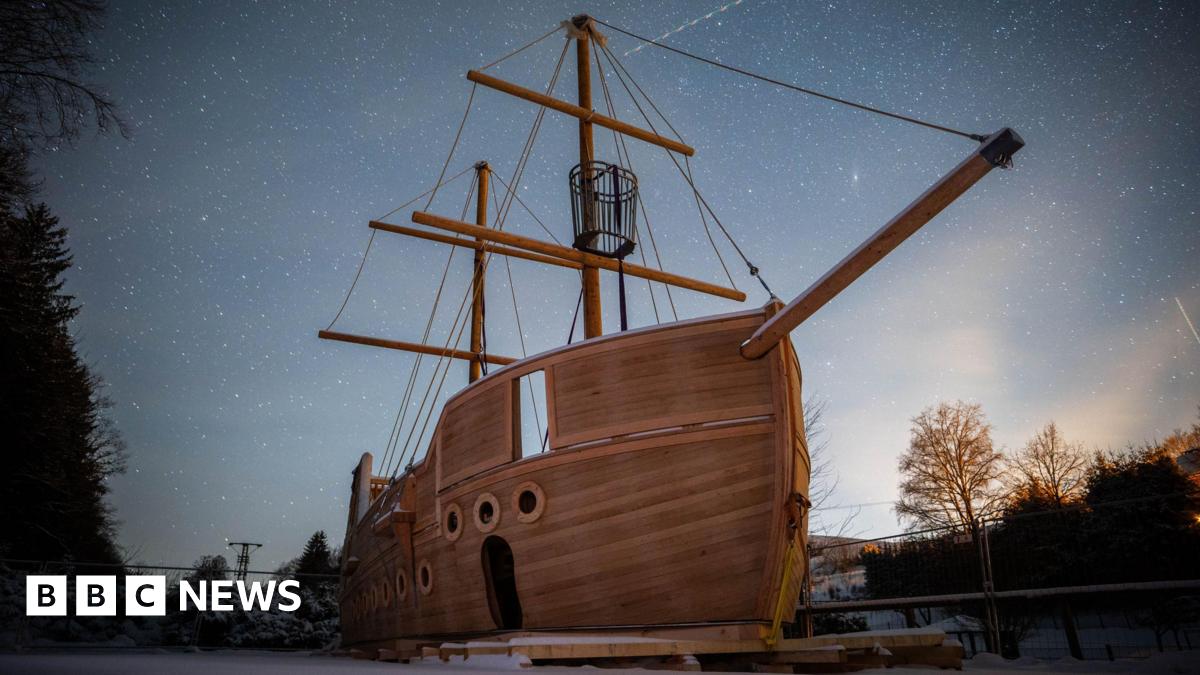 Millie Bobby Brown and David Harbour in character as Eleven and Jim Hopper. Eleven wears red shorts over grey leggings and a grey jumper. Jim Hopper wears a black leather jacket, a black beanie hat and dark trousers.They are speaking to director Frank Darabont on the set of Stranger Things. There is a vintage advertisement posted in the background with debrief on the ground.