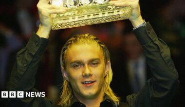 The snooker player Paul Hunter, lifting a trophy aloft out of shot. His blond hair is braided and he is wearing a black shirt.